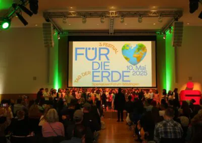 Ein großes Publikum beobachtet Kinder, die auf der Bühne vor einer Leinwand mit der Aufschrift Für die Erde, einer herzförmigen Erde und dem Datum 10. Mai 2025 im SparkassenForum Böblingen, mit grüner Beleuchtung an den Wänden.