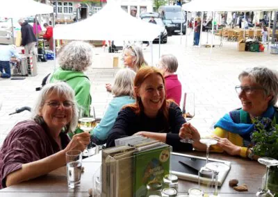 Drei Frauen sitzen an einem Tisch im Freien, lächeln und halten Getränke in der Hand, auf dem Tisch stehen Bücher und Pflanzen. Im Hintergrund sind andere Menschen und Marktstände auf einem belebten Platz zu sehen.