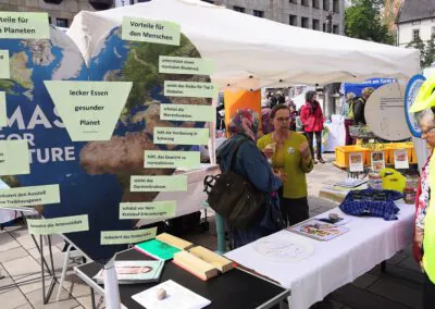 Ein Klimabewusstseinsstand bei einer Veranstaltung im Freien zeigt Informationsplakate in deutscher Sprache über die Vorteile von Klimaschutzmaßnahmen. Die Menschen diskutieren miteinander, und auf Tischen liegen verschiedene Broschüren und Modelle aus.