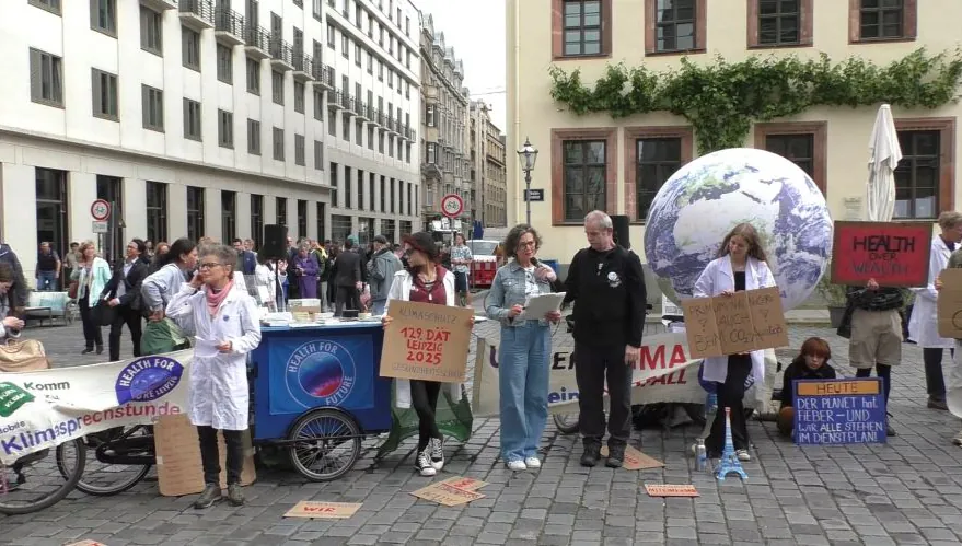 Eine Gruppe von Menschen steht bei einem Klimaprotest im Freien und hält Schilder und Transparente. Einige tragen Laborkittel und eine Person ist als die Erde verkleidet. Zu den Requisiten gehören ein großer Globus und ein Tisch, der mit Materialien für den Klimaschutz bedeckt ist.