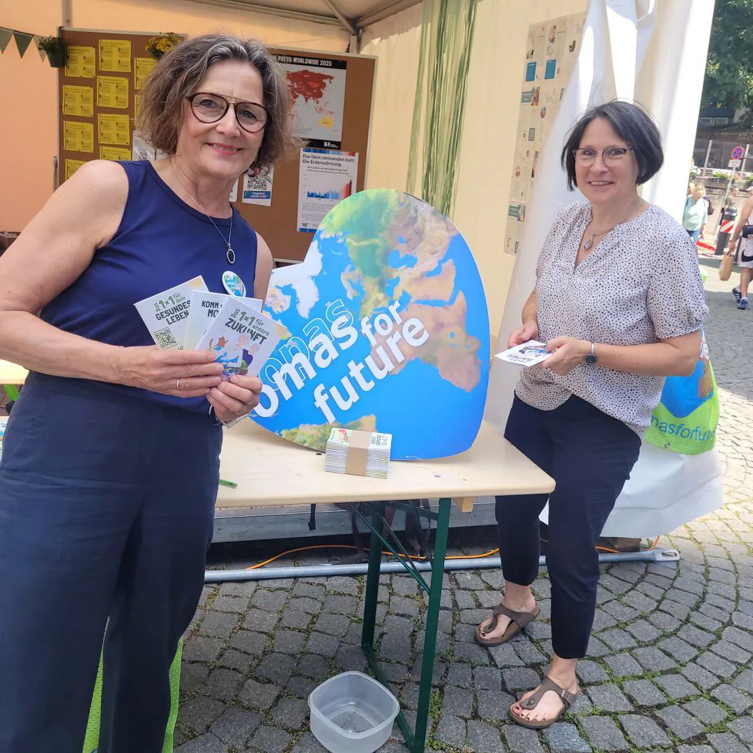 Zwei Frauen stehen an einem Stand im Freien mit einem großen erdförmigen Schild, auf dem "Omas for Future" steht. Sie lächeln, halten Flugblätter in der Hand, und auf dem Tisch liegt Informationsmaterial aus. Im Hintergrund eine Straße mit Kopfsteinpflaster.