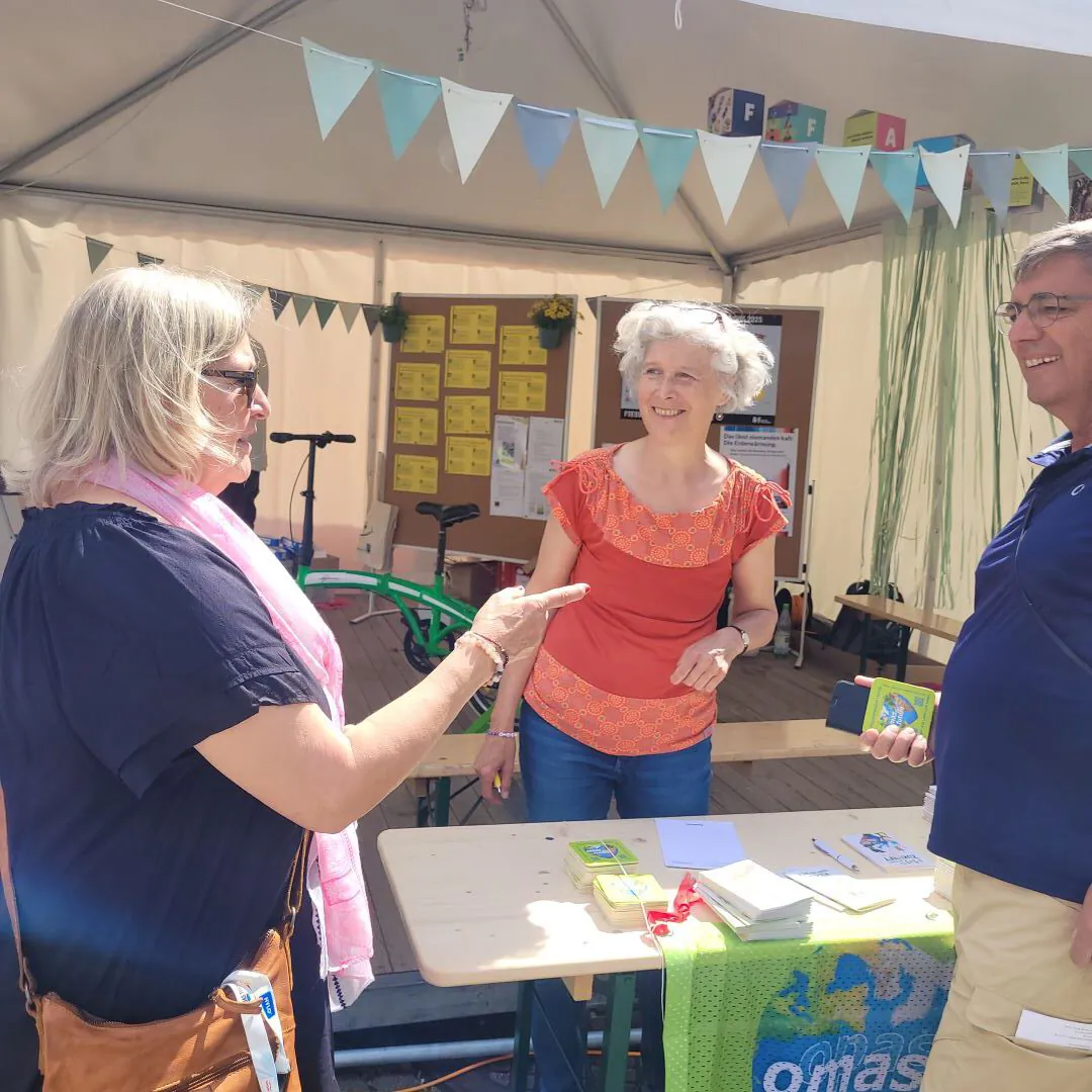 Drei Erwachsene stehen an einem Außenstand mit Informationsbroschüren und unterhalten sich. Zwei Frauen und ein Mann lächeln unter einem mit blauen Wimpeln geschmückten Zelt. Ein Tisch mit Broschüren und ein grünes Fahrrad sind zu sehen.