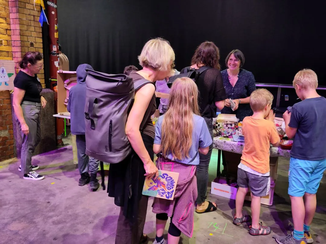 Eine Gruppe von Erwachsenen und Kindern versammelt sich bei einer Indoor-Veranstaltung um einen Tisch mit Bastel- und Kunstzubehör. Eine Frau hält ein Kinderbuch, während die anderen die Gegenstände auf dem Tisch erkunden.