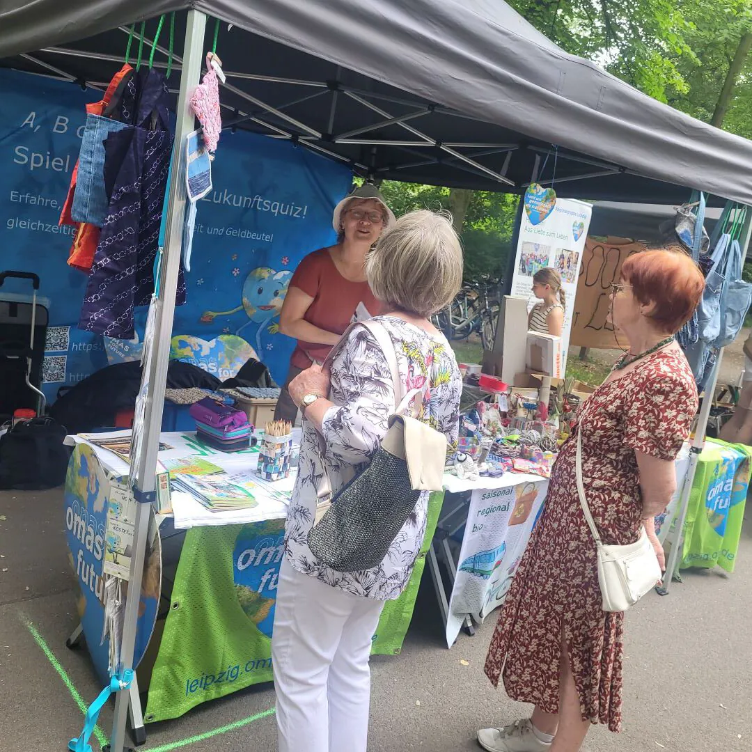 Drei Frauen stehen an einem überdachten Stand im Freien und unterhalten sich. Der Stand zeigt bunte Informationsmaterialien, Kunsthandwerk und Banner zum Thema Nachhaltigkeit. Im Hintergrund sind Bäume und andere Menschen zu sehen.