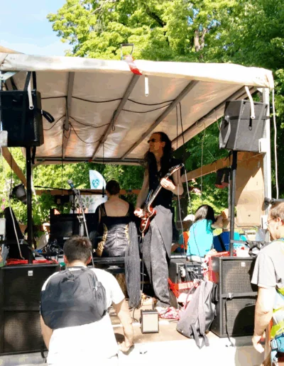 Eine Band tritt auf einer überdachten Bühne im Freien auf, auf der ein Musiker Gitarre spielt. Hinter der Bühne schauen Menschen zu. Bäume und Grünpflanzen füllen den Hintergrund im hellen Sonnenlicht.