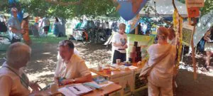 Muenchner O4F Klimadult 2025 1©O4F - Omas for Future Menschen versammeln sich auf einer Freiluftmesse mit Informationsständen unter Bäumen. An Tischen, die mit Bannern und farbenfrohen Auslagen geschmückt sind, unterhalten sich die Menschen und verteilen Informationsmaterial. Das Sonnenlicht fällt durch die Blätter und schafft eine lebendige Atmosphäre.