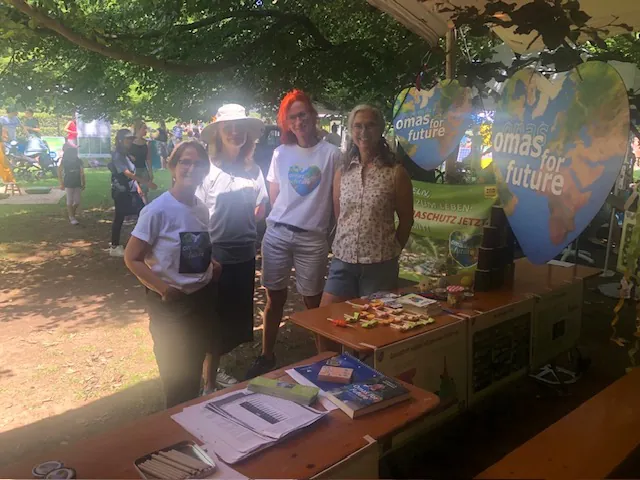 Vier Frauen stehen lächelnd an einem Stand im Freien unter einem Baum, mit herzförmigen Omas for Future-Schildern, Flyern, Snacks und Informationsmaterial auf dem Tisch. Im Hintergrund sind andere Menschen und Grünflächen zu sehen.
