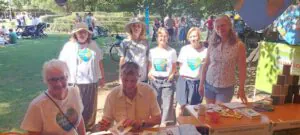 Muenchner O4F Klimadult 2025 3©O4F - Omas for Future Eine Gruppe von sieben Personen steht und sitzt an einem Stand mit Bastelmaterial im Freien. Die meisten tragen T-Shirts mit herzförmigen Erdmotiven. Im sonnigen Park im Hintergrund sind Menschen und Spaziergänger zu sehen.