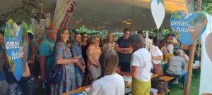 Muenchner O4F Klimadult 2025 Stadtrat©O4F - Omas for Future Eine Gruppe von Menschen steht unter einem großen Veranstaltungszelt und ist um einen Tisch mit Informationsmaterial versammelt. Herzförmige Schilder mit der Aufschrift "omas" für "Zukunft" und der Darstellung der Erde deuten auf eine umwelt- oder klimabezogene Veranstaltung hin.