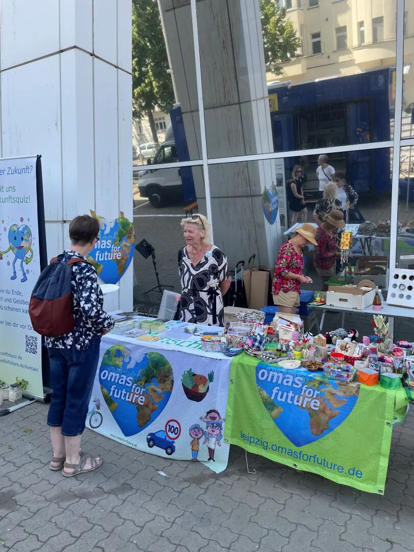 Zwei Frauen stehen hinter einem Tisch mit Umweltartikeln und Schildern mit der Aufschrift "Omas for Future" an einem Straßenstand im Freien. Eine Frau mit einem Rucksack spricht zu ihnen. Im Hintergrund sind ein großes Gebäude und reflektierende Fenster zu sehen.