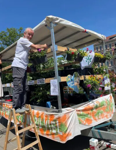 Ein älterer Mann steht auf einer mit Topfblumen und bunten Bannern bedeckten Plattform während einer sonnigen Veranstaltung im Freien. An der Struktur sind Schilder mit Illustrationen und Text angebracht. Im Hintergrund sind Bäume und Gebäude zu sehen.