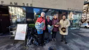 WilderMuell©O4FBB SIFI - Omas for Future Vier ältere Frauen stehen lächelnd vor einem Café und halten Schilder in Form von Herzen mit der Aufschrift OMAS FOR FUTURE. Sie sind umgeben von Müllsäcken und Gartengeräten, die auf eine gemeinschaftliche Aufräumaktion hindeuten.