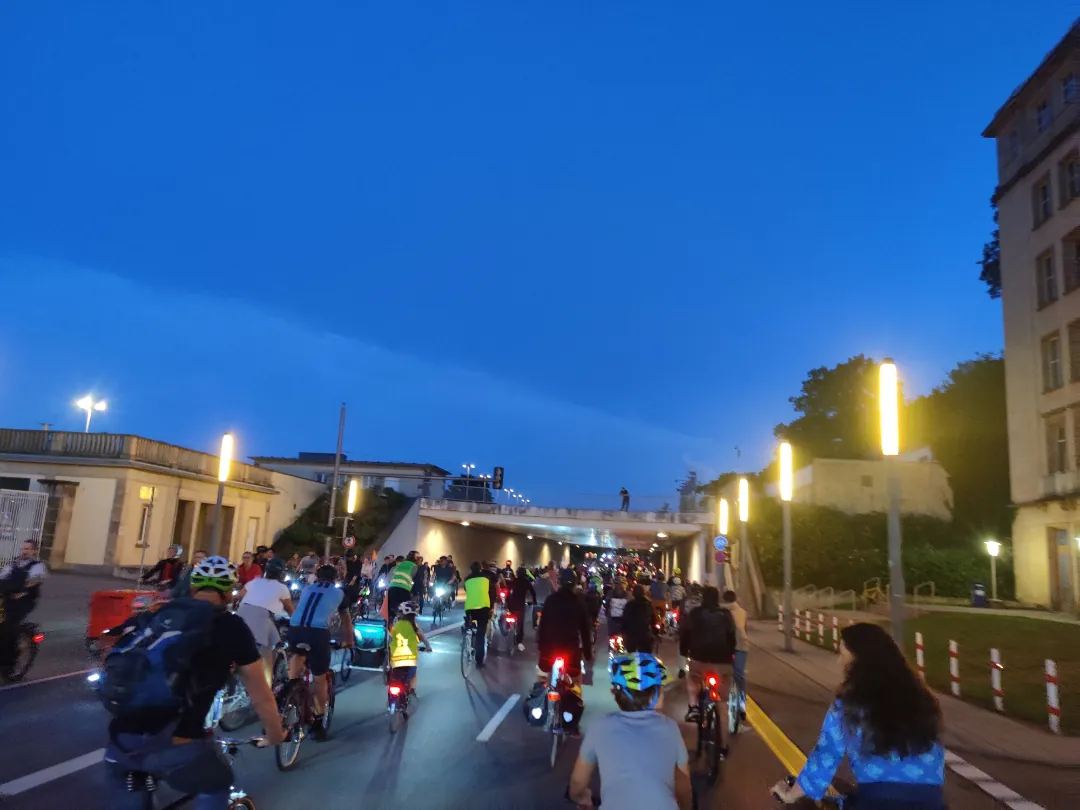 Eine große Gruppe von Radfahrern fährt in der Abenddämmerung gemeinsam auf einer Stadtstraße unter einer Brücke hindurch. Die Straßenlaternen leuchten, und einige Radfahrer tragen Helme und Rucksäcke. Der Himmel ist tiefblau mit Gebäuden und Bäumen im Hintergrund.