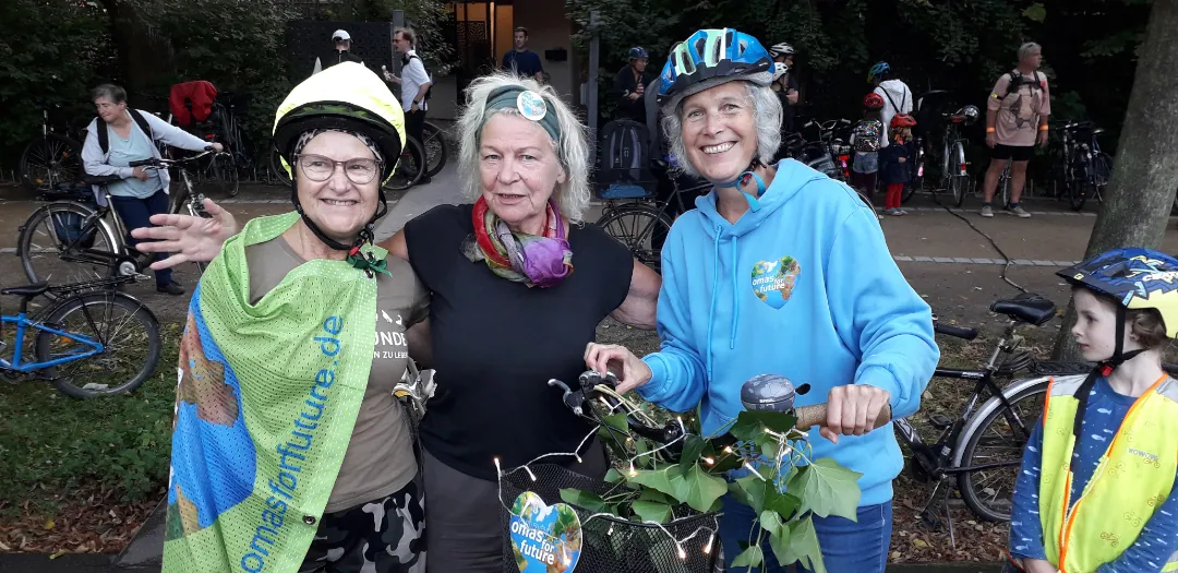 Drei lächelnde Frauen, zwei mit Fahrradhelmen und eine mit einem Schal, stehen bei einer Fahrradveranstaltung zusammen. Eine trägt einen grünen Umhang mit Text, eine andere hält ein geschmücktes Fahrrad. Im Hintergrund sind Menschen und Fahrräder zu sehen, und ein Kind mit einem Helm schaut zu.