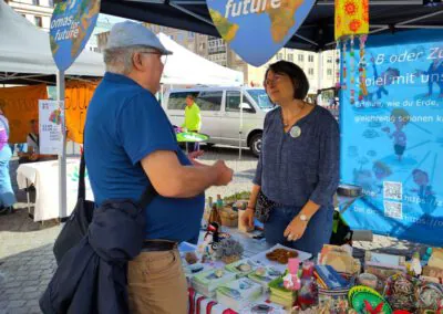 Ein Mann unterhält sich mit einer Frau an einem Umweltstand, der umweltfreundliche Artikel und Informationsmaterial ausstellt. Darüber hängen Banner mit der Aufschrift "Omas for future", und im Hintergrund sind weitere Marktstände zu sehen.