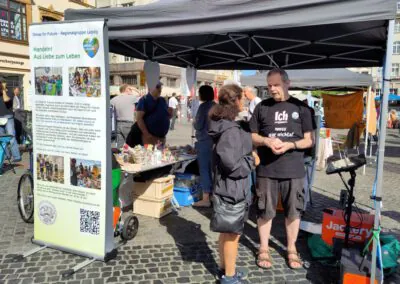 Zwei Personen unterhalten sich an einem Stand im Freien unter einem Vordach. In der Nähe steht ein großes Schild mit Fotos und deutschem Text. Auf Tischen sind verschiedene Gegenstände ausgestellt, und im Hintergrund sind mehrere Personen auf einem Platz mit Kopfsteinpflaster zu sehen.