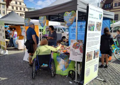 Menschen unterhalten sich an einem Marktstand mit Bannern zum Thema Nachhaltigkeit. Eine Frau im Rollstuhl steht vor dem Tisch, und andere stehen in der Nähe. Im Hintergrund sind Stände, Zelte und ein historisches Gebäude zu sehen.