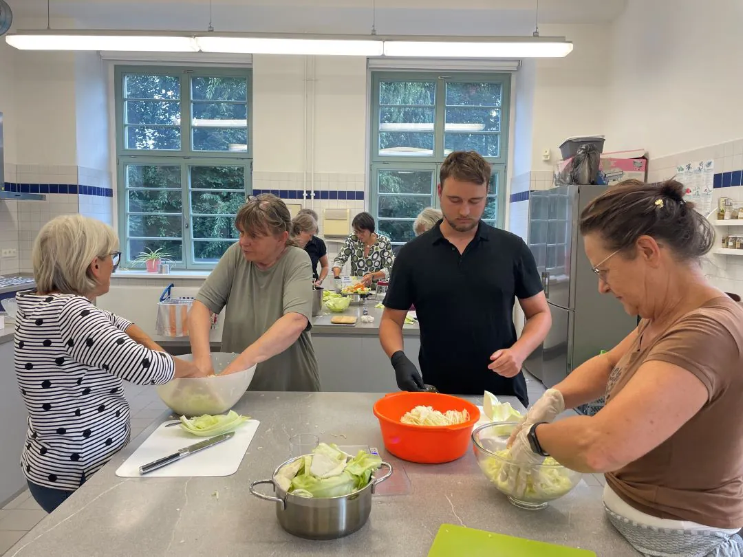 Vier Personen stehen mit Handschuhen um einen Küchentisch herum und bereiten das Essen mit Schüsseln und gehacktem Kohl zu. Im Hintergrund arbeiten weitere Personen. Die Küche ist hell mit großen Fenstern und weiß gekachelten Wänden.