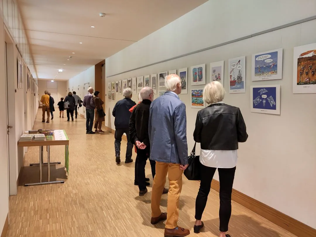 Menschen gehen einen hellen Flur entlang und betrachten gerahmte Kunstwerke, die in einer Ausstellung an den Wänden hängen. Die meisten Betrachter sind ältere Erwachsene, einige stehen und betrachten die Bilder genau. Der Boden ist aus Holz und auf der linken Seite stehen Tische.