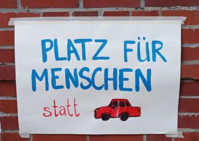 Auf einem handgemalten Schild, das an eine Backsteinmauer geklebt wurde, steht in blauen und roten Buchstaben "Platz für Menschen statt", darunter die Zeichnung eines roten Autos.