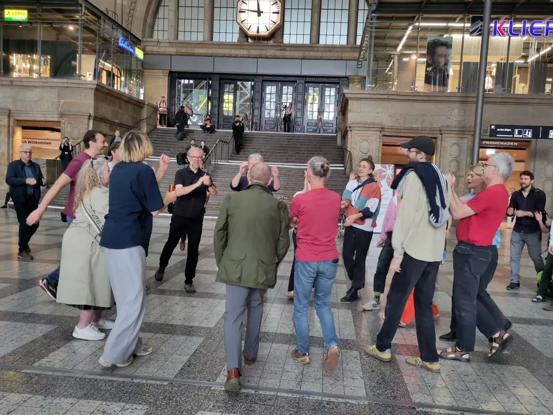 Eine Gruppe von Menschen steht im Kreis, klatscht und unterhält sich angeregt in einem großen Bahnhof mit einer großen Treppe und einer Uhr im Hintergrund. Passanten und Schaulustige beobachten die Szene.