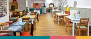 IMG 5349 - Omas for Future Ein gemütliches Café mit Holzböden, bunten Stühlen und Tischen mit Speisekarten und Blumen. Die Wände sind mit Fotos und Kunstwerken dekoriert, und die Theke ist im hinteren Teil des Raumes zu sehen.