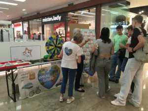 Menschen versammeln sich um einen "Omas for Future"-Informationsstand in einem Einkaufszentrum mit einem Preisrad und Postern über Nachhaltigkeit. Die Käufer interagieren mit Freiwilligen neben einem Display, das für umweltfreundliche Praktiken wirbt.