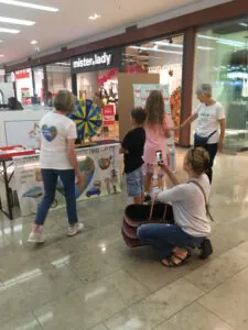 Eine Frau kniet nieder, um ein Foto von zwei Kindern zu machen, die vor einem bunten Spinnrad an einem Stand in einem Einkaufszentrum stehen, assistiert von zwei Frauen in weißen T-Shirts. Im Hintergrund sind Geschäfte und Schilder zu sehen.
