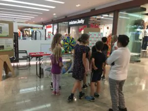 Eine Gruppe von Kindern und ein Erwachsener versammeln sich um ein buntes Spinnrad an einem Stand in einem Einkaufszentrum, mit Auslagen und Postern im Hintergrund.