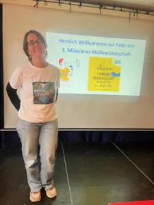 Eine lächelnde Frau in legerer Kleidung und mit Brille steht vor einem Bildschirm, auf dem eine Willkommensfolie für die 3. Sie trägt ein T-Shirt mit der Aufschrift "omas for future".