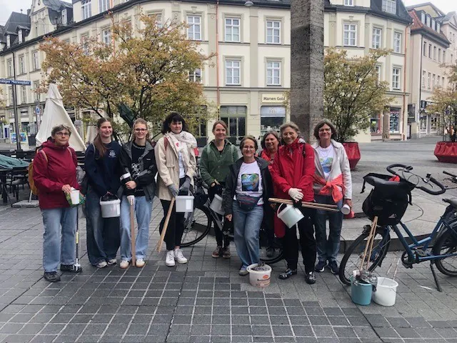 Eine Gruppe von neun Personen steht mit Eimern und Werkzeugen in der Hand im Freien auf einer Straße in Bad Kreuznach. Im Hintergrund sind Bäume mit Herbstlaub, Gebäude mit Geschäften, einige Fahrräder und ein paar Topfpflanzen zu sehen.