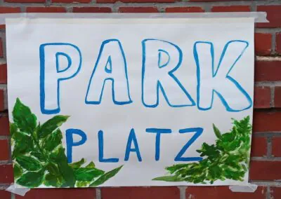 Auf einem handgemalten Schild, das an einer Backsteinmauer befestigt ist, steht in blauen Buchstaben PARK PLATZ, mit grün bemalten Blättern an den unteren Ecken.