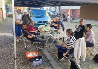 Eine Gruppe von Menschen sitzt um einen Tisch im Freien unter einem Vordach und teilt Essen und Getränke. Einige sitzen auf Campingstühlen, andere stehen und lächeln, was eine freundliche und lockere Atmosphäre auf einer Kopfsteinpflasterstraße schafft.