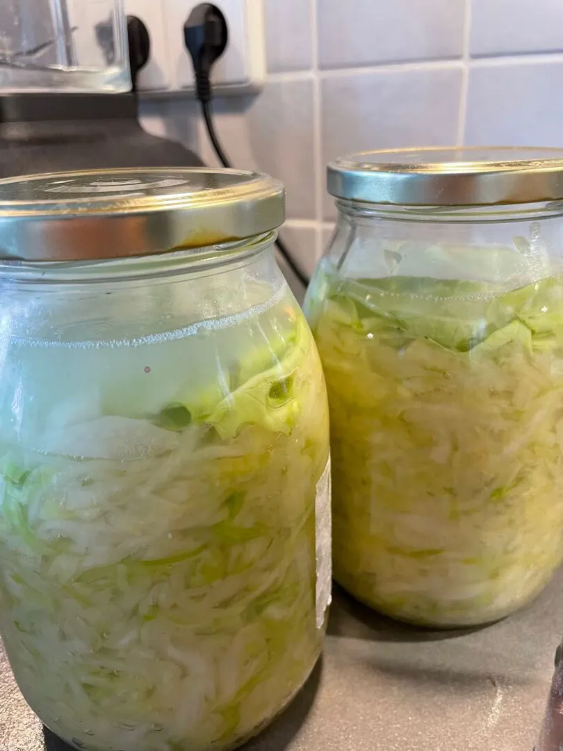 Zwei Gläser mit in Scheiben geschnittenem und in Flüssigkeit getauchtem Kohl, wahrscheinlich selbstgemachtes Sauerkraut oder Essiggurken, stehen auf einem Küchentisch in der Nähe einer gekachelten Wand und einer Steckdose.