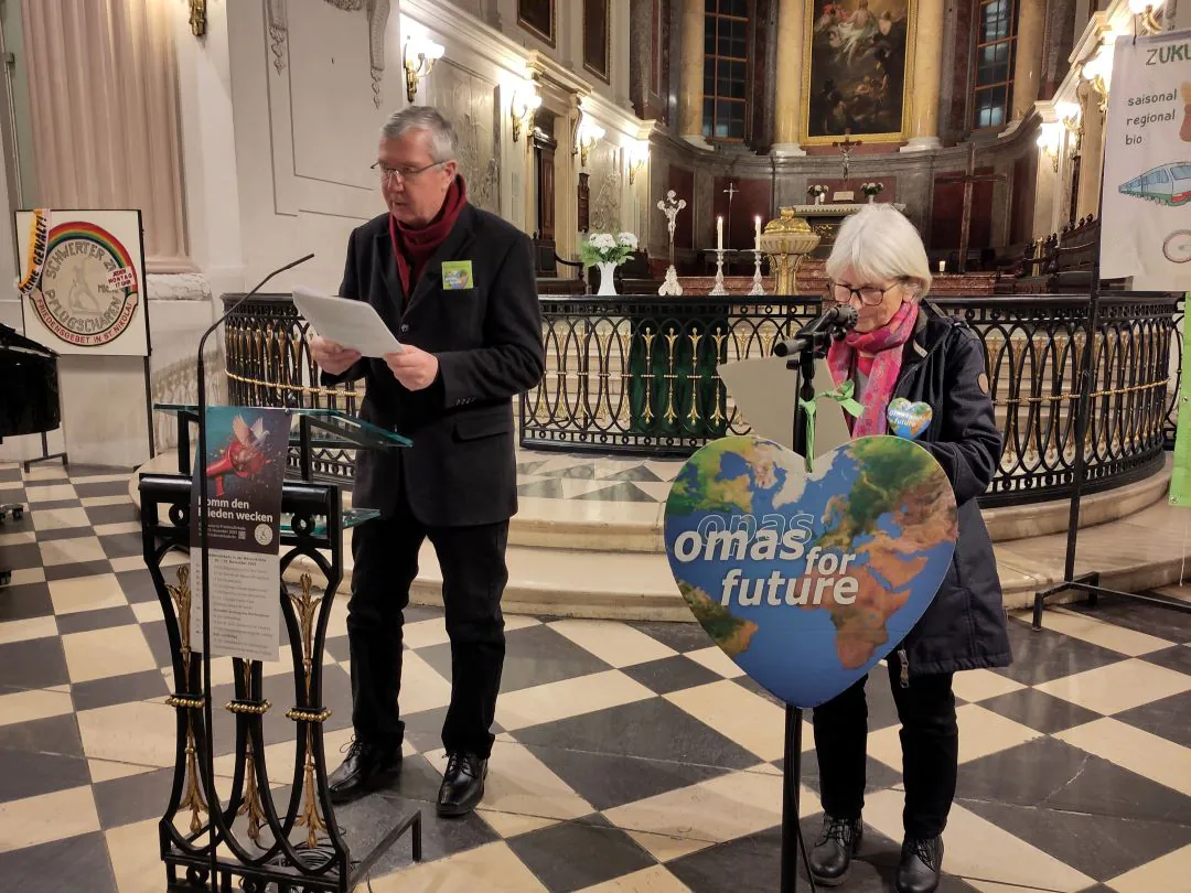 Zwei ältere Erwachsene stehen auf einem Podium in einer Kirche und sprechen auf einer Veranstaltung. Auf dem Podium der Frau steht ein herzförmiges Schild mit der Aufschrift "Omas for Future". Im Hintergrund sind Kerzen, Kirchendekoration und ein Klavier zu sehen.