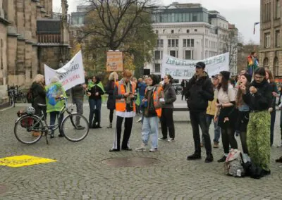 Eine Gruppe von Menschen versammelt sich zu einem Protest auf einem städtischen Platz, einige halten Schilder und Transparente. Einige tragen orangefarbene Sicherheitswesten, und ein Fahrrad steht an der Seite. Im Hintergrund sind Gebäude und Bäume zu sehen.