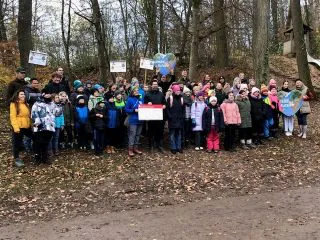 Eine große Gruppe von Kindern und Erwachsenen steht draußen in einem Waldstück zusammen, einige halten Schilder und herzförmige Plakate in der Hand. Alle sind warm angezogen, und der Boden ist mit abgefallenen Blättern bedeckt.