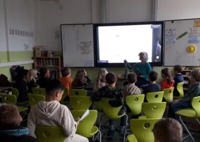 Ein Lehrer sitzt vorne in einem Klassenzimmer und arbeitet mit einem digitalen Whiteboard, während eine Gruppe junger Schüler auf grünen Stühlen sitzt und aufmerksam zuhört. Der Raum ist gut beleuchtet und mit Unterrichtsmaterialien ausgestattet.