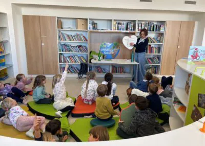 Eine Lehrerin steht an einer Tafel, hält ein herzförmiges Schild in der Hand und wendet sich an eine Gruppe kleiner Kinder, die auf bunten Kissen in einer Bibliothek mit vielen Bücherregalen sitzen. Einige Kinder haben ihre Hände erhoben.