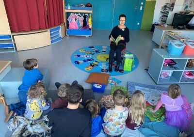 Eine Frau sitzt auf einem Stuhl und liest einer Gruppe kleiner Kinder, die in einem Halbkreis um sie herum auf dem Boden sitzen, in einem farbenfrohen Klassenzimmer ein Buch vor. Die Kinder hören aufmerksam zu, einige sitzen auf Kissen oder Decken.