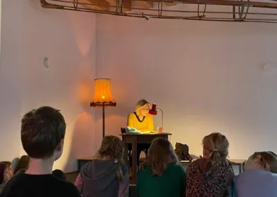 Eine Frau sitzt an einem Schreibtisch unter einer Schreibtischlampe und liest einer Gruppe von Kindern vor, die vor ihr auf dem Boden sitzen. Eine alte Stehlampe leuchtet warm neben ihr in einem ansonsten schummrigen, minimalistischen Raum.