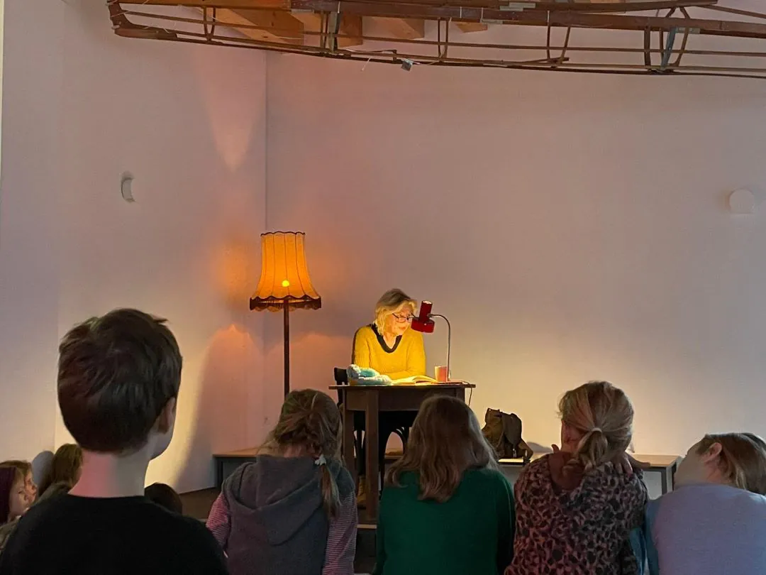 Eine Frau sitzt an einem Schreibtisch unter einer Schreibtischlampe und liest einer Gruppe von Kindern vor, die vor ihr auf dem Boden sitzen. Eine alte Stehlampe leuchtet warm neben ihr in einem ansonsten schummrigen, minimalistischen Raum.