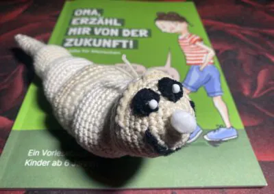 Eine gehäkelte Raupe mit großen Augen liegt auf einem grünen Kinderbuch mit dem Titel "Oma, erzähl mir von der Zukunft!", auf dessen Cover eine Frau und eine Raupe abgebildet sind.