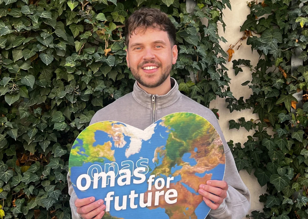 Ein lächelnder junger Mann steht vor belaubtem Efeu und hält ein herzförmiges Schild mit einer Weltkarte und der Aufschrift "omas for future" in der Hand.