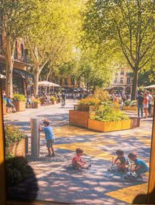 Superblock Stadt fuer alle Muenchen 2026 ©O4F - Omas for Future Ein sonnenbeschienener Stadtplatz mit Menschen, die in Straßencafés sitzen, grüne Bäume säumen den Platz. Im Vordergrund spielen Kinder auf dem bemalten Pflaster, ein Kind an einem Wasserbrunnen, während andere mit Kreide zeichnen.