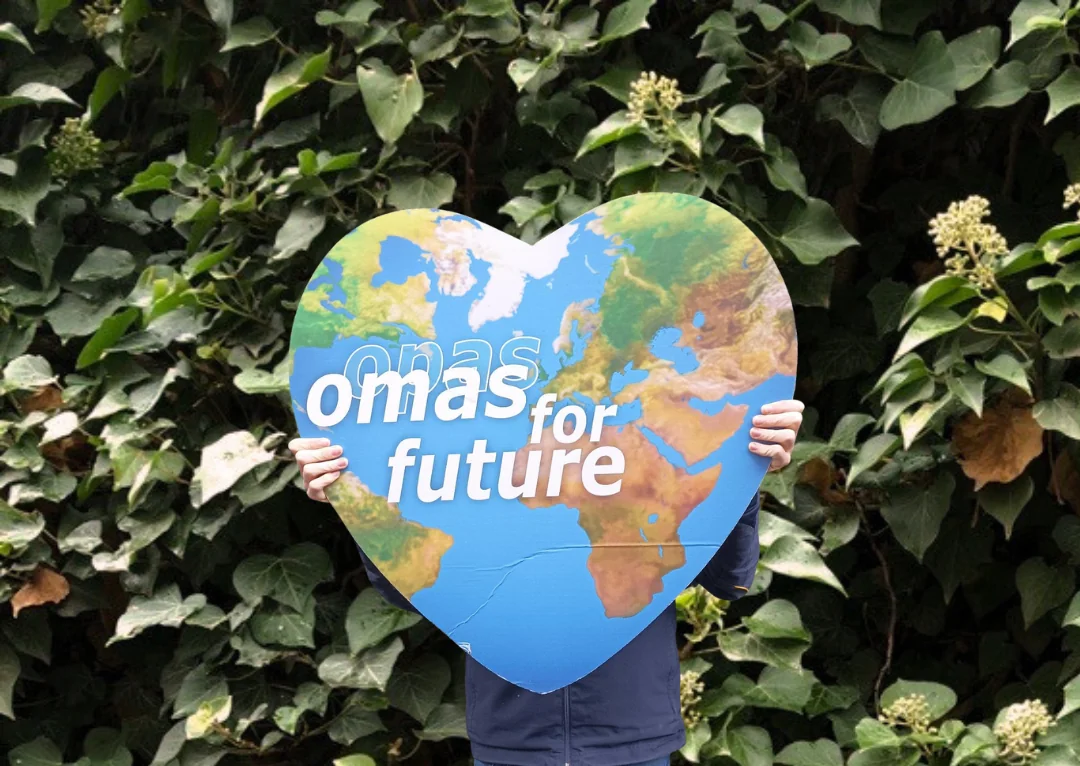 Ein Teammitglied hält ein großes, herzförmiges Schild mit einer Karte von Europa und Teilen von Afrika und Asien. Auf dem Schild steht omas for future vor einem grünen Hintergrund, wobei das Gesicht der Person hinter dem Schild verborgen ist.