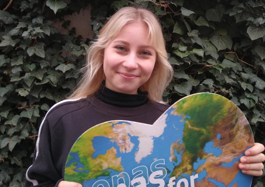 Eine junge Person mit langen blonden Haaren, die einen schwarzen Pullover trägt, lächelt und hält ein herzförmiges Schild mit einer Weltkarte. Grüne Efeublätter bedecken den Hintergrund.