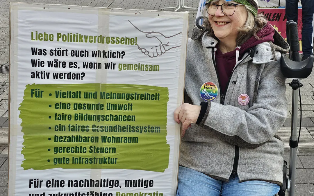 Eine ältere Frau mit Brille und Mütze sitzt in einem Rollstuhl und lächelt neben einem Protestschild. Auf dem Schild stehen Forderungen nach Demokratie, Vielfalt, Umwelt, gerechter Bildung, bezahlbarem Wohnraum und besserer Infrastruktur.