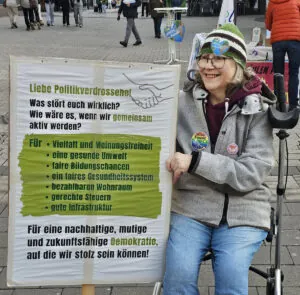 ©OFF KH Machen statt meckern 1 - Omas for Future Eine ältere Frau mit Brille und Mütze sitzt in einem Rollstuhl und lächelt neben einem Protestschild. Auf dem Schild stehen Forderungen nach Demokratie, Vielfalt, Umwelt, gerechter Bildung, bezahlbarem Wohnraum und besserer Infrastruktur.