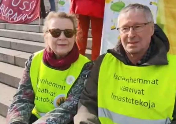 Zwei Erwachsene, die leuchtend gelbe Sicherheitswesten mit finnischem Text tragen, sitzen auf einer Treppe bei einer öffentlichen Demonstration oder Kundgebung. Eine Person trägt eine Sonnenbrille; beide schauen in die Kamera. Im Hintergrund sind Menschen und Transparente zu sehen.